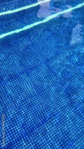 top view of the clear blue water in a swimming pool with tiled floor and gentle ripples on the surface under the sun. High quality 4k footage, vertical
