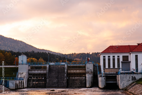 Hydroelectric dam and water control building by reservoir at sunset with forested hills, colorful sky and industrial infrastructure,