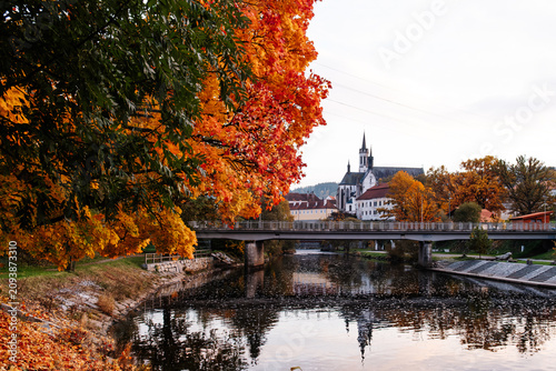 Historic monastery and church by river with bridge, colorful autumn foliage and reflections on calm water, concept of European travel, architecture and cultural heritage.