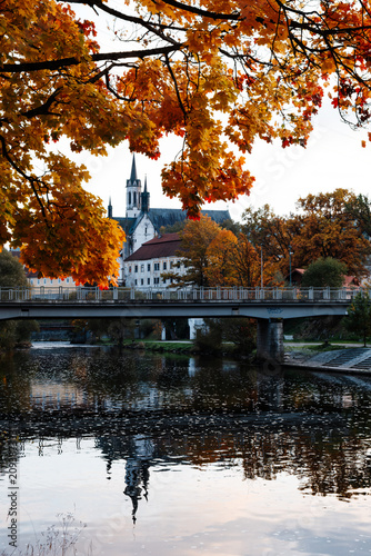 Historic monastery and church reflected in calm river water with bridge and colorful autumn foliage