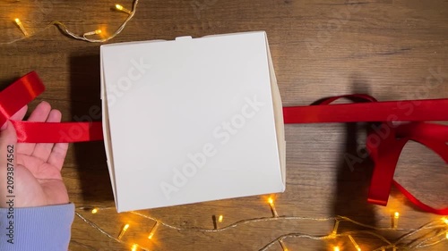 4K video close up. Top view. White woman ties red satin bow on white box with Merry Christmas, Happy New Year 2027, birthday gift. Garland with yellow gold glow light on table. Winter holiday concept