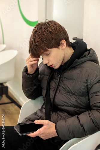 Принти на полотні Teenage boy in dark puffer jacket absorbed in smartphone while sitting in waitin