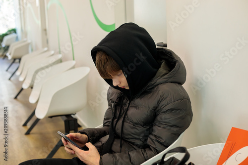 Konstfotografi Teenage boy in dark puffer jacket absorbed in smartphone while sitting in waitin