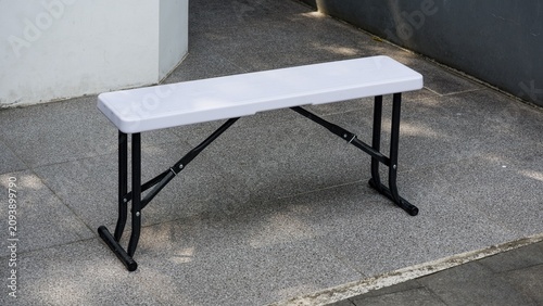 An empty white plastic folding bench with black metal legs sitting on an outdoor stone patio floor.