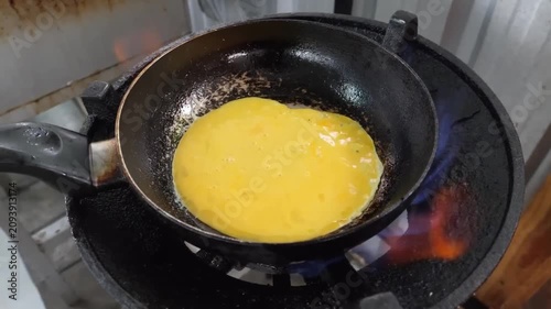A detailed shot capturing the culinary process of pouring a bright yellow egg mixture from a ceramic bowl into a seasoned pan over a blue gas flame. This high-energy footage highlights street food coo