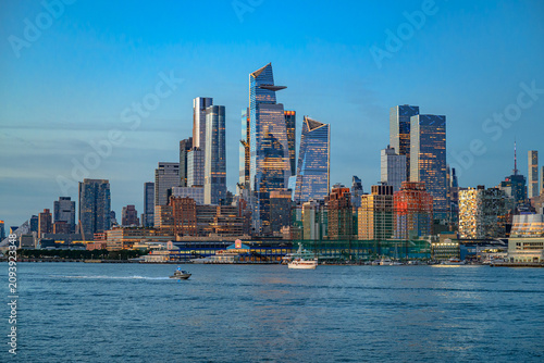 Hudson Yards skyline across Hudson River at dusk. Glass skyscrapers rise above waterfront piers as boats cross the river under a blue evening sky.