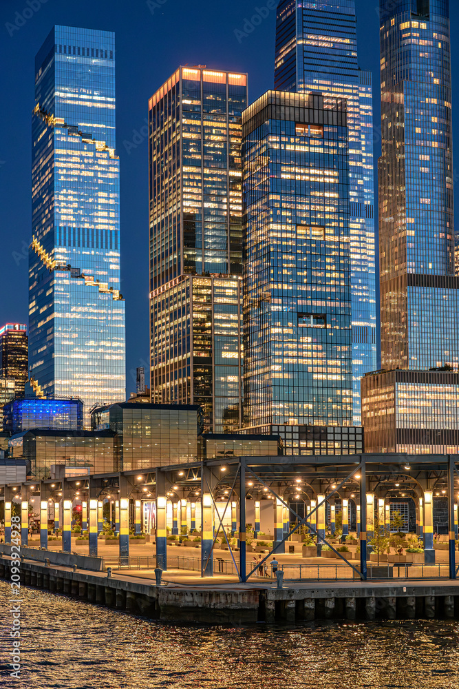 Fototapeta premium Pier 76 beneath illuminated Hudson Yards office towers. Covered waterfront structure with lit columns borders the river below dense glass skyscrapers at night.