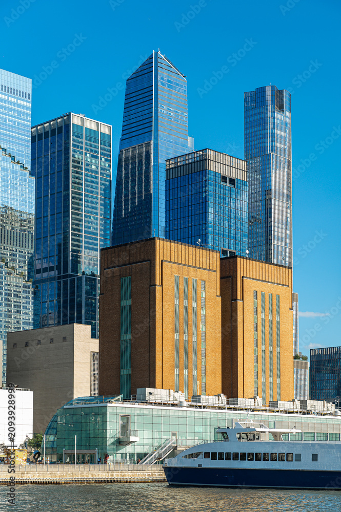 Fototapeta premium Hudson Yards glass towers behind Manhattan waterfront building. Vertical composition shows layered skyscrapers, brick architecture, a ferry, and clear blue sky.