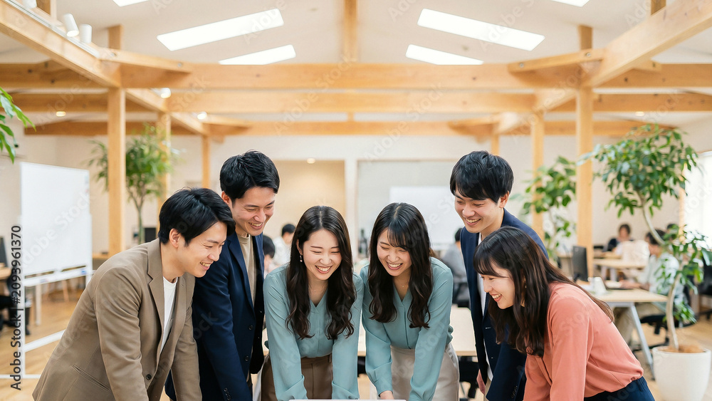 Obraz premium 木造梁のある明るいオフィスで窓辺の木製デスクを囲む六人の同僚がきゃっきゃと笑い合いながらタブレット画面を覗き込む活気ある議論を楽しむ様子を明るい日差しの中で描写しています。