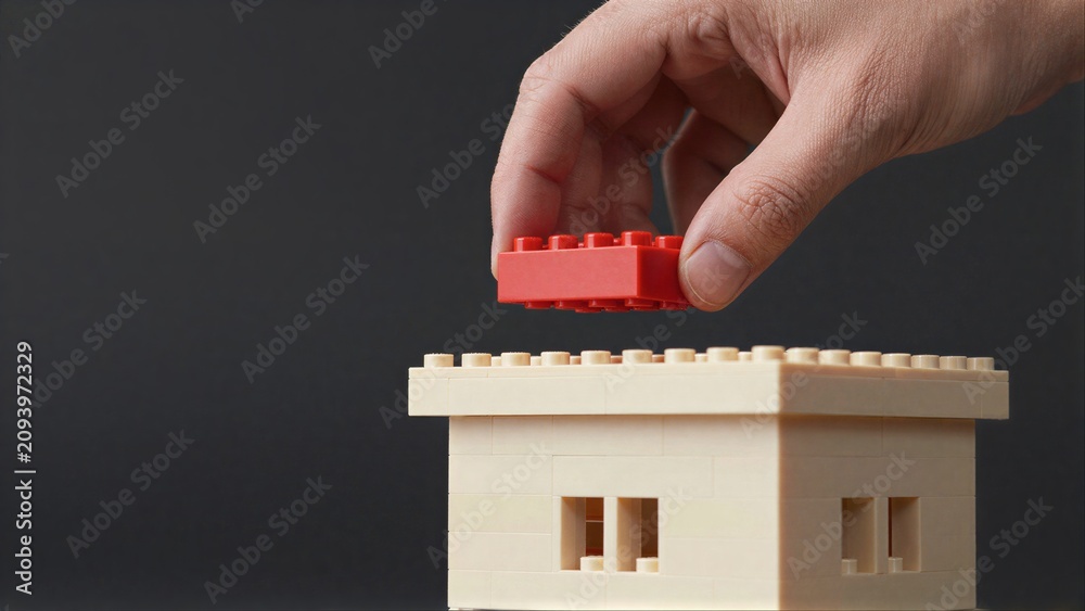 Fototapeta premium Hand places red LEGO brick atop beige structure, symbolizing growth or construction.