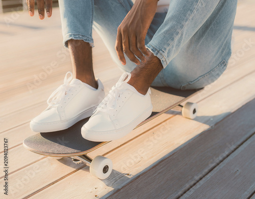 Person sitting on skateboard with white sneakers on wooden deck daytime close-up view photography