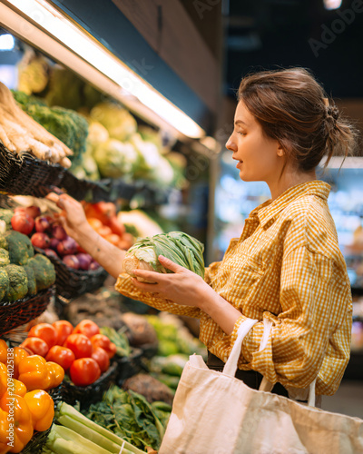 Young woman comparing fresh seasonal vegetables inside a premium grocery produce section, reusable shopping bag, colorful natural food display, authentic healthy shopping lifestyle