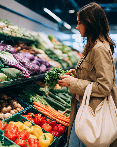Young woman comparing fresh seasonal vegetables inside a premium grocery produce section, reusable shopping bag, colorful natural food display, authentic healthy shopping lifestyle