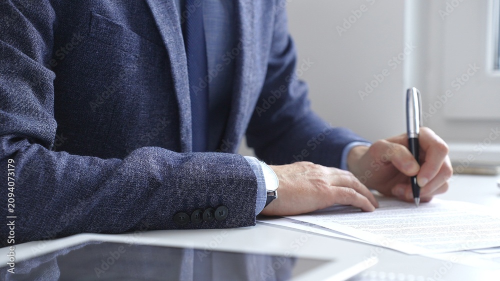 Obraz premium Professional businessman signing contract with silver pen, wearing tailored blue suit, business documents and digital tablet nearby. Business people