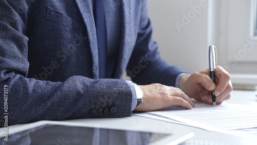Professional businessman signing contract with silver pen, wearing tailored blue suit, business documents and digital tablet nearby. Business people