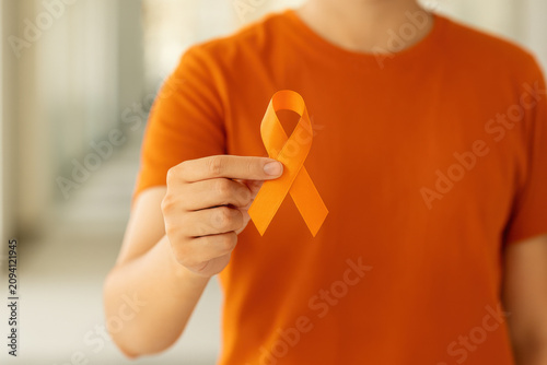Person wearing orange shirt holding orange leukemia awareness ribbon toward camera with blurred indoor background and soft natural lighting