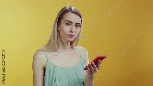 A young woman is engaged with her smartphone while standing against a vibrant yellow background