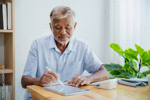 Elderly Asian man using digital tablet and stylus for health monitoring and takes notes at home.