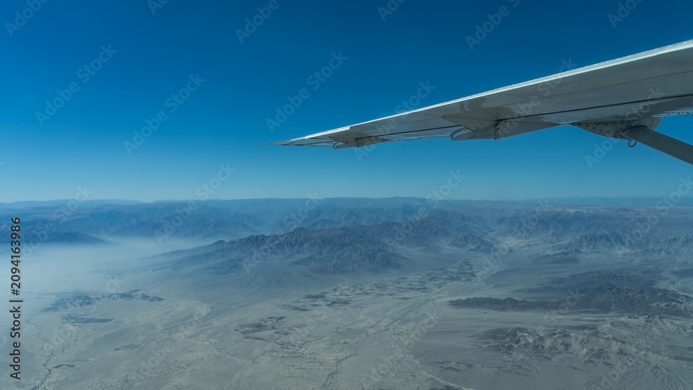 Naklejka premium Flying over the desert. An airplane wing on a blue sky background. Below there are sand dunes, hills, mountains without vegetation, winding riverbeds of dried rivers. Peru. The Nazca Plateau.