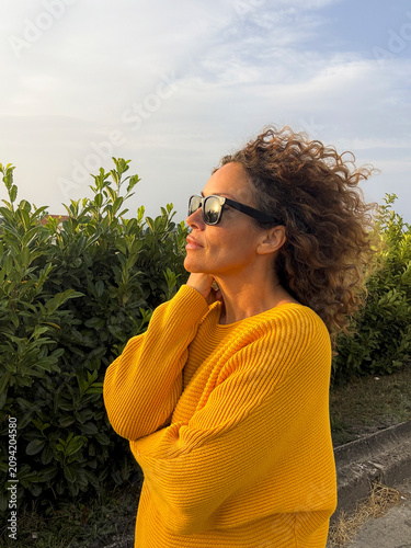 Fotografie Woman in a yellow sweater wearing sunglasses stands outdoors by green hedges at