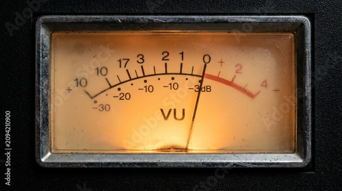 Close-up of a vintage VU meter with a red needle on a black background, warm orange lighting, and a metal frame.