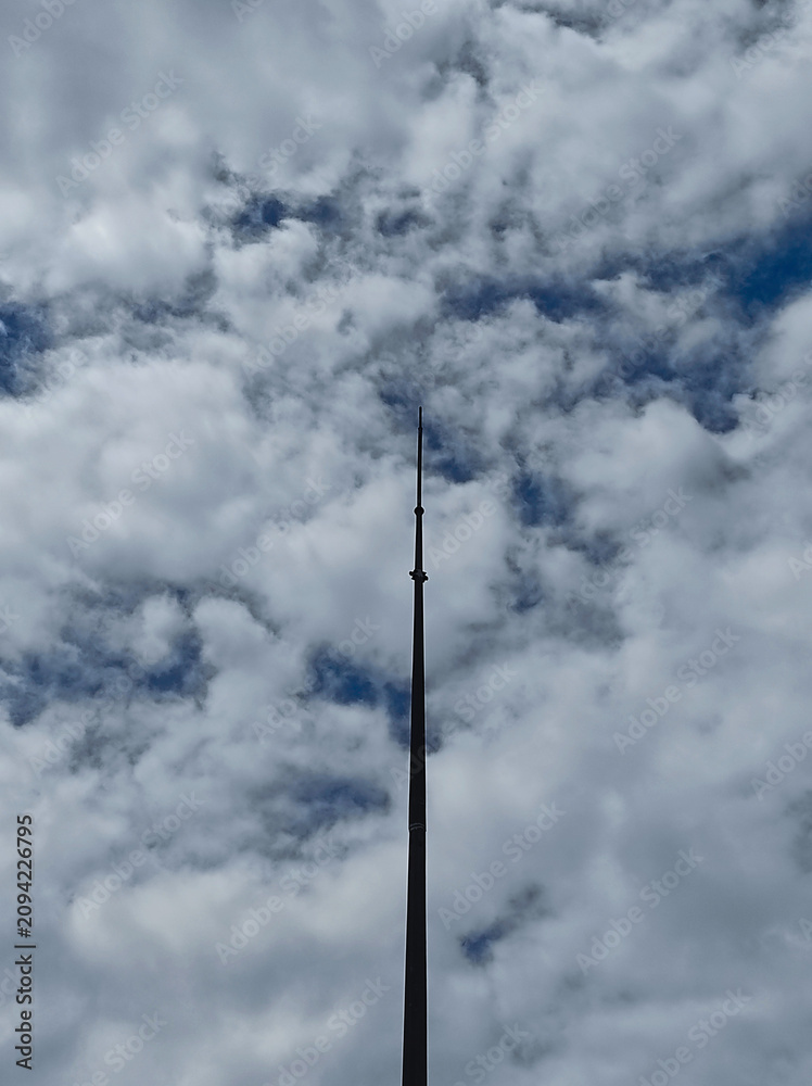 Obraz premium A towering lightning rod pierces a dramatic, cloudy sky streaked with deep blue. A minimalist, vertical shot from a low angle shows the contrast between the man-made structure and nature.