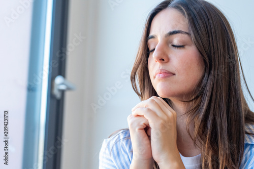 Fotografie Young woman praying with eyes closed near a window, peaceful and contemplative, expressing faith, hope and gratitude in a quiet indoor portrait