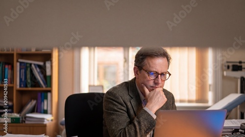 A thoughtful middle-aged man with glasses wearing a tweed jacket sits at his desk, intently focused on a laptop screen, with bookshelves filled with study materials in the background.