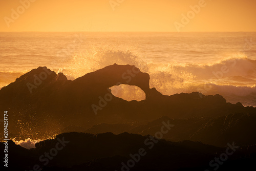 The golden orange light of sunrise shines through splashing surf, highlighting the hole caused by a small natural rock arch on the coast at the harbour entrance to Narooma, New South Wales, Australia.