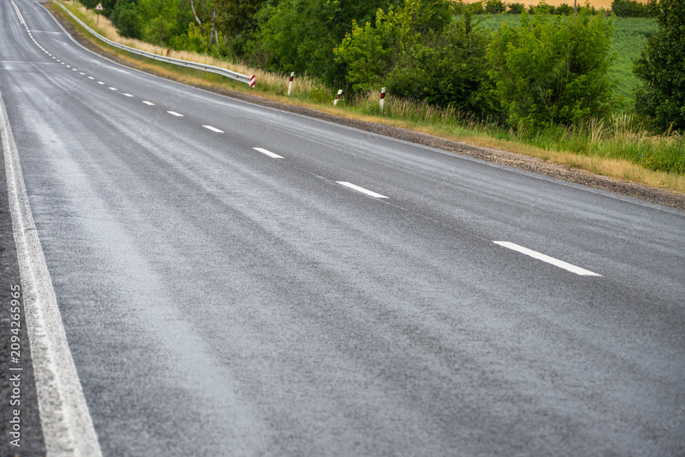 Obraz premium An empty asphalt highway with dashed white dividing lines gently curves through a rural landscape bordered by lush green trees