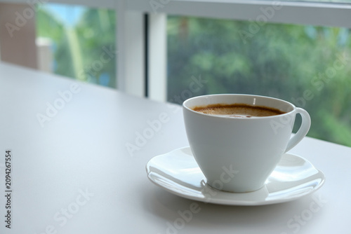 Coffee cup saucer white table window foam drink morning beverage in minimalist kitchen with blurred green garden view