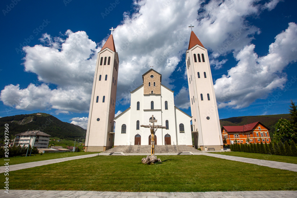 Naklejka premium Church of the Holy Family with Two Towers and Crucifix Sculpture under Dramatic Sky in Kupres Bosnia and Herzegovina