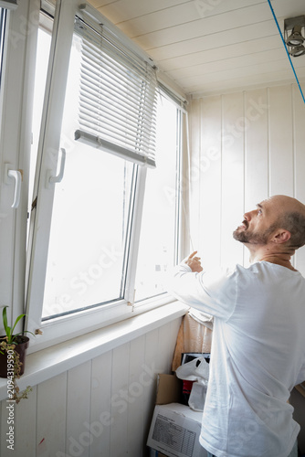 Middle Aged Man Adjusting White Venetian Blinds on Plastic Window on Balcony