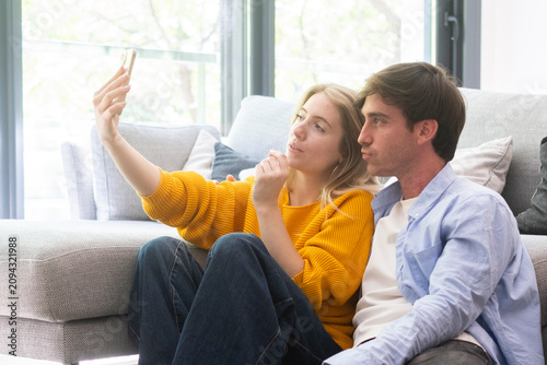 Bild auf Leinwand Young couple sitting on a living room floor taking a playful selfie together, relaxed and cozy at home, smiling and making faces while leaning against a sofa in natural daylight