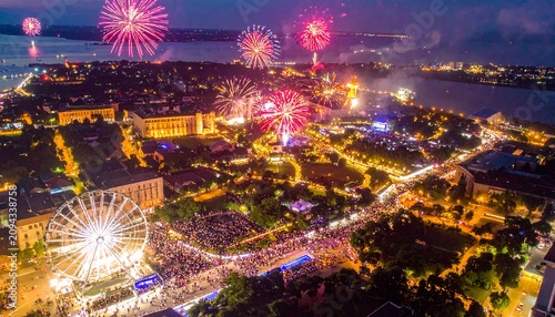 Aerial view of a city celebrating with fireworks over illuminated buildings, water, and a fair