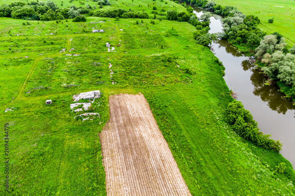 Obraz premium Aerial view of overgrown abandoned farmland with decaying greenhouse ruins, remnants of old agricultural structures set in a vibrant green meadow beside a calm winding river