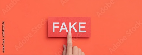 Hand pointing at red paper sign with white text reading fake on solid orange background representing misinformation and digital deception concepts