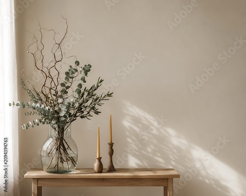 Glass vase with eucalyptus branches and dried twigs next to two taper candles on a wooden console table against a beige wall