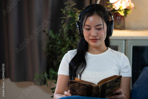 A peaceful Asian woman wears wireless headphones while reading a vintage book on the sofa at night.