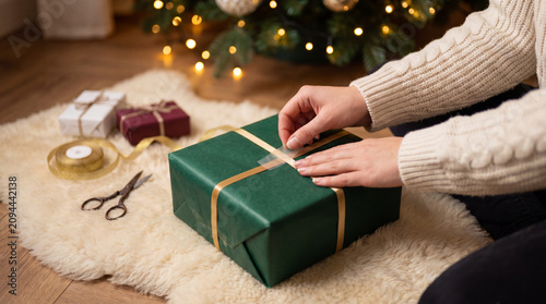 Hands wrapping a Christmas gift with gold ribbon beneath a decorated tree