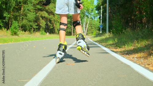 Child girl in casual clothes roller skating on road in forest on sunny summer day.