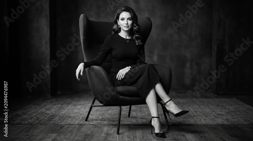 Elegant businesswoman in black dress sitting in wingback chair, cinematic black and white portrait of a confident female leader in luxury studio