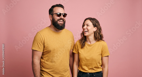 Laughing young couple wearing yellow t-shirts and jeans standing together against a pink background