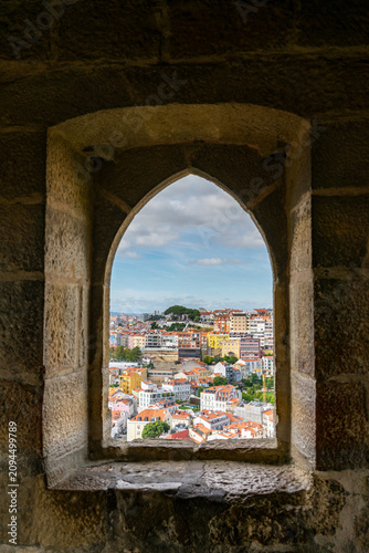 Konstfotografi A thick, rustic stone arched window captures a scenic view of the densely packed, colorful historic city of Lisbon, Portugal, rolling over a distant hill under a bright sky with scattered clouds