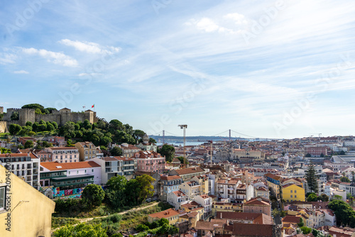 Obraz na płótnie The sprawling cityscape of Lisbon, Portugal, with the historic Castelo de São Jorge on a prominent green hill to the left, overlooking a valley of terracotta-roofed buildings
