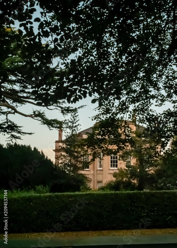 A stately, multi-story Georgian-style country house stands nestled among dense, green foliage and trees at dusk, viewed from behind a neatly trimmed hedge that frames the architectural details.