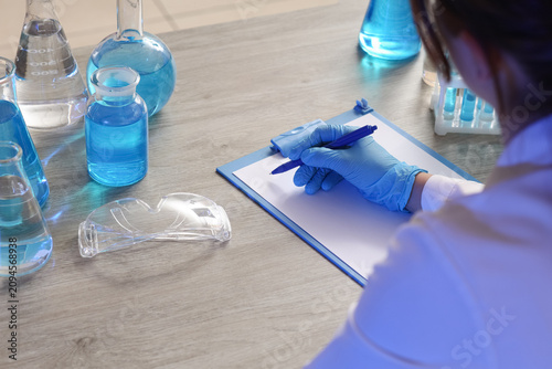Scientist in blue gloves writing on a clipboard with laboratory glassware and...