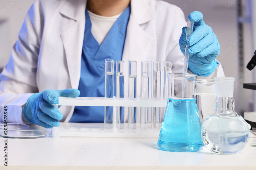 Fototapeta premium Female scientist taking water sample from flask on stand to test tubes at table in laboratory, closeup