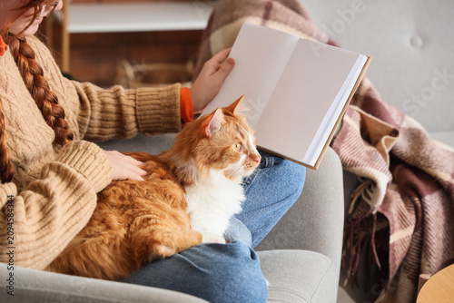 Teenage redhead girl with cute cat reading book in armchair at home