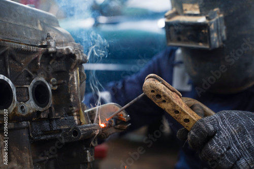 Close-up of a professional welder wearing safety gear, welding a metal car engine part with sparks flying in an industrial workshop.